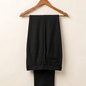 May include: A pair of solid black trousers featuring a structured waistband with belt loops, a clean front design, and a single visible welt pocket on the back. The trousers are displayed hanging on a wooden clothes hanger against a neutral background.