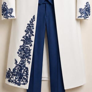 May include: A white long coat featuring intricate dark blue floral embroidery along the front panel and cuffs, paired with solid dark blue trousers. The coat has a structured open front design, while the trousers feature a clean, tailored finish.