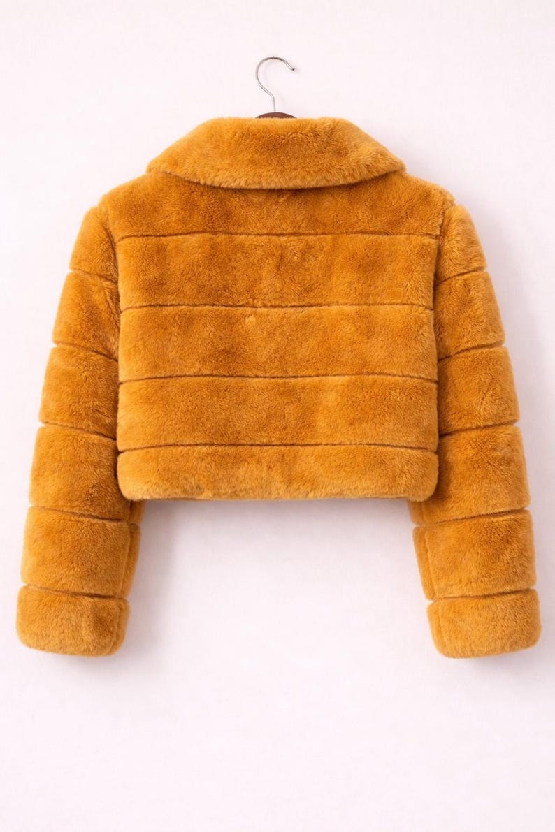 May include: Mustard-colored faux fur jacket with a collar and horizontal stripe design. The cropped jacket is hanging on a white background. The jacket has long sleeves.