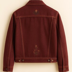 May include: A burgundy denim jacket with floral embroidery on the back. The jacket features a classic collar, button closures, and two chest pockets. The floral design includes shades of yellow, orange, and green.