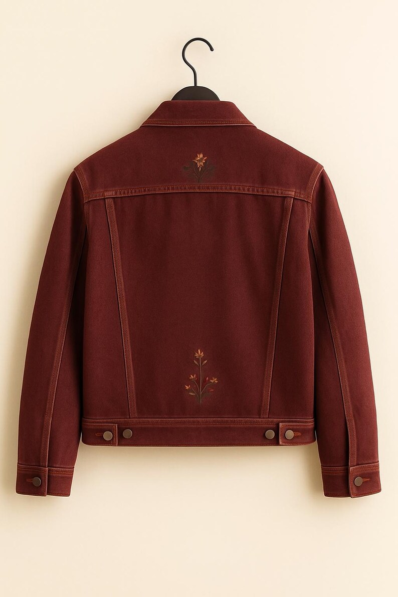 May include: A burgundy denim jacket with floral embroidery on the back. The jacket features a classic collar, button closures, and two chest pockets. The floral design includes shades of yellow, orange, and green.