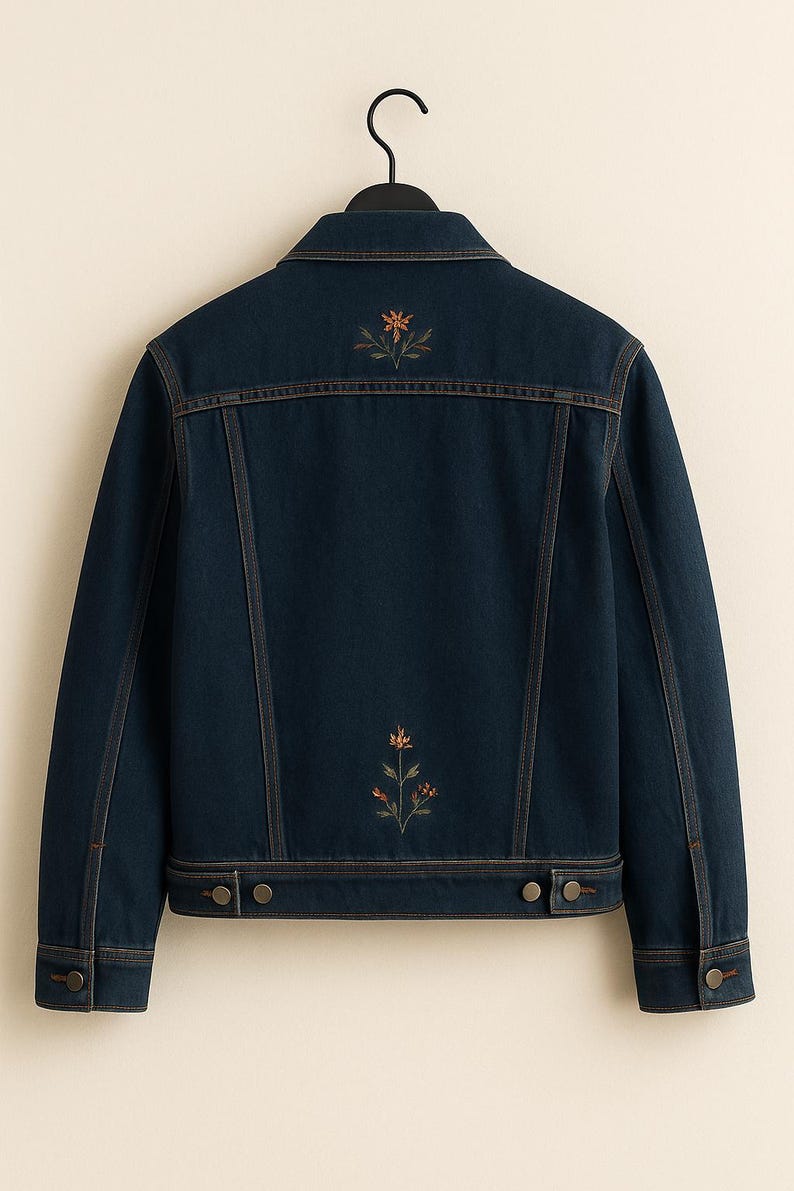 May include: A dark blue denim jacket featuring a classic collar, long sleeves with buttoned cuffs, and two adjustable button tabs at the hem. The back displays two small, embroidered floral motifs in orange and green thread centered vertically.