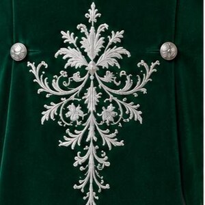 Puede incluir: Una prenda de terciopelo verde intenso con un gran patr&oacute;n de bordado plateado intrincado en el centro. El dise&ntilde;o consta de motivos florales y de hojas sim&eacute;tricos y en espiral. Se aprecian dos botones redondos de metal plateado.