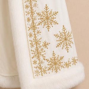 May include: A white cloak featuring a thick white faux fur trim along the edges and a vertical panel decorated with intricate gold embroidered snowflake patterns of varying sizes, creating a detailed winter-themed design on the fabric.