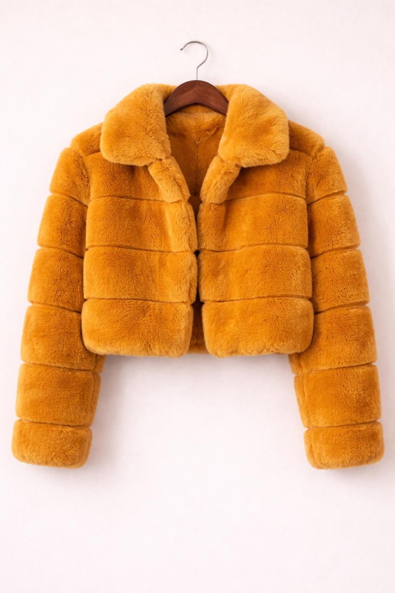 May include: Mustard-colored faux fur jacket with a collared neckline and horizontal stripe design. The cropped jacket is hanging on a wooden hanger against a white background. The jacket has long sleeves.