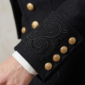 May include: A black coat featuring intricate black embroidery on the cuffs and gold-toned metal buttons. The sleeve cuff displays a detailed swirling pattern, while the front of the coat is adorned with a vertical row of matching gold buttons.