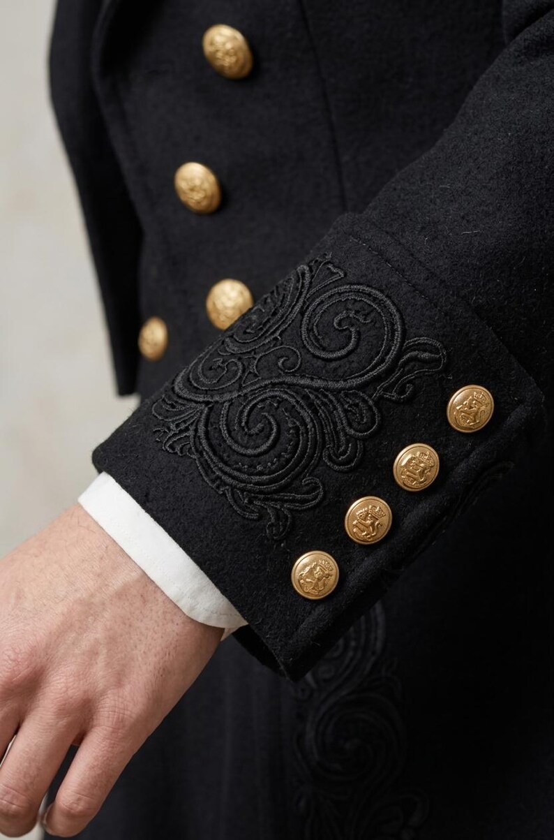 May include: A black coat featuring intricate black embroidery on the cuffs and gold-toned metal buttons. The sleeve cuff displays a detailed swirling pattern, while the front of the coat is adorned with a vertical row of matching gold buttons.