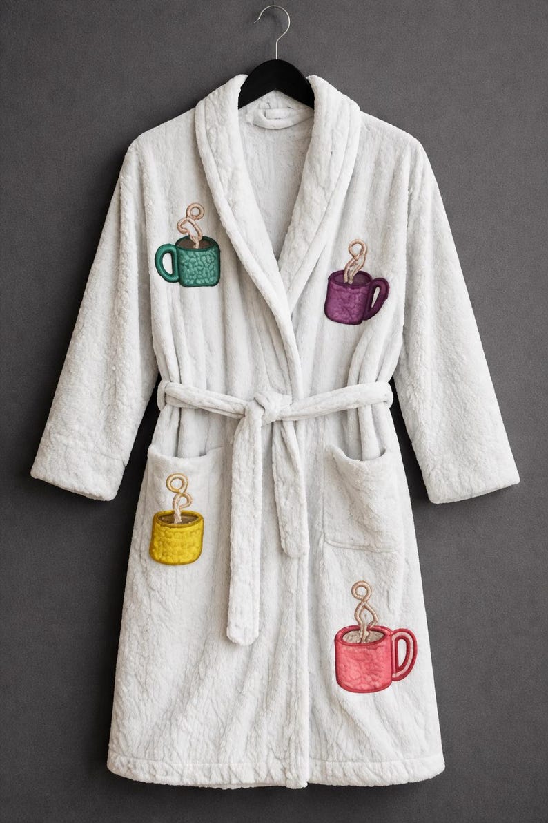 Puede incluir: Bata de felpa blanca con dise&ntilde;os bordados de tazas de caf&eacute; en verde, morado, amarillo y rojo. La bata tiene cuello chal, cintur&oacute;n en la cintura y dos bolsillos delanteros. Cuelga de una percha negra.