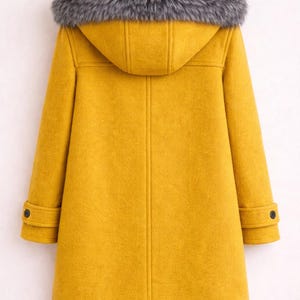 Puede incluir: Un abrigo amarillo mostaza con capucha y ribete de piel sint&eacute;tica gris. El abrigo tiene un dise&ntilde;o cl&aacute;sico de abrigo duffle con cierres de palanca y pu&ntilde;os abotonados. El abrigo est&aacute; colgado de una percha de madera.