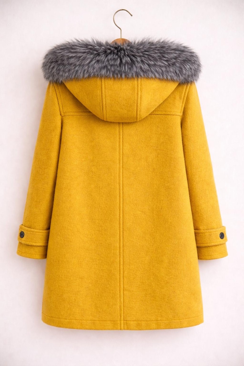 Puede incluir: Un abrigo amarillo mostaza con capucha y ribete de piel sint&eacute;tica gris. El abrigo tiene un dise&ntilde;o cl&aacute;sico de abrigo duffle con cierres de palanca y pu&ntilde;os abotonados. El abrigo est&aacute; colgado de una percha de madera.