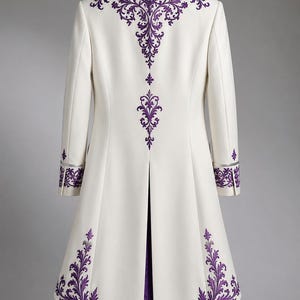 May include: A long white coat featuring intricate purple embroidery along the collar, back, cuffs, and hem. The garment has a structured silhouette with a center back vent, long sleeves, and a high collar, displayed on a wooden hanger.