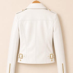 May include: A white faux leather jacket with gold-coloured hardware, including zips on the sleeves and decorative buckles at the waist. The jacket is hanging on a wooden hanger, showing its back with detailed stitching.
