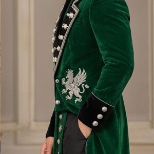 May include: A long-length emerald green velvet coat featuring a black velvet lapel, silver metallic embroidery of a griffin on the side, and silver buttons along the front closure and cuffs, paired with plain black trousers.
