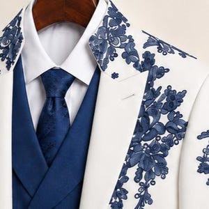 May include: A white blazer featuring dark blue floral embroidery on the lapels, worn over a solid blue vest and a crisp white collared shirt with a matching dark blue patterned necktie. The ensemble displays a structured, formal appearance.