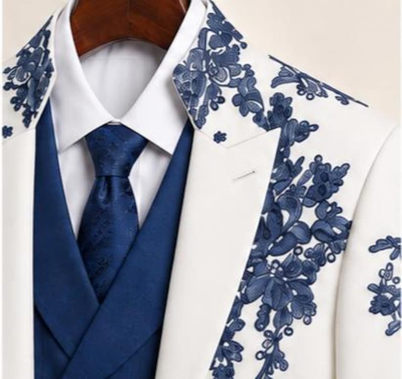 May include: A white blazer featuring dark blue floral embroidery on the lapels, worn over a solid blue vest and a crisp white collared shirt with a matching dark blue patterned necktie. The ensemble displays a structured, formal appearance.