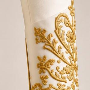 May include: A white cylindrical sleeve cuff featuring raised gold-colored relief ornamentation. The decorative pattern consists of intricate swirling floral and leaf motifs that wrap around the exterior surface, finished with a thin gold border trim.
