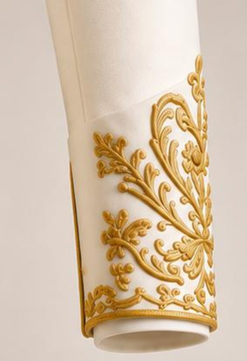 May include: A white cylindrical sleeve cuff featuring raised gold-colored relief ornamentation. The decorative pattern consists of intricate swirling floral and leaf motifs that wrap around the exterior surface, finished with a thin gold border trim.