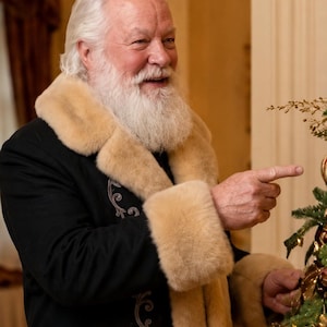 May include: A person with white hair and a full white beard wearing a black coat with a thick beige faux fur collar and cuffs. The coat features intricate silver embroidery along the front opening. The person is pointing toward a decorated tree.