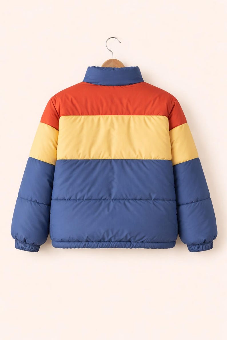 May include: A color-blocked puffer jacket in red, yellow, and blue. The jacket has a high collar and elastic cuffs. The jacket is hanging on a wooden hanger. The jacket is a casual outerwear garment.