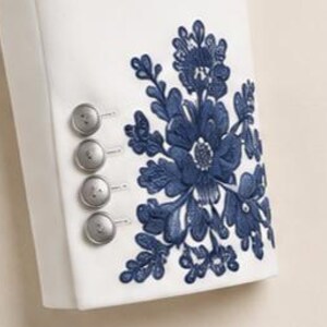 May include: A white sleeve featuring a vertical row of four round silver buttons and a large, intricate dark blue floral embroidery pattern. The embroidery consists of detailed petals and leaves, positioned next to the buttons on the fabric.