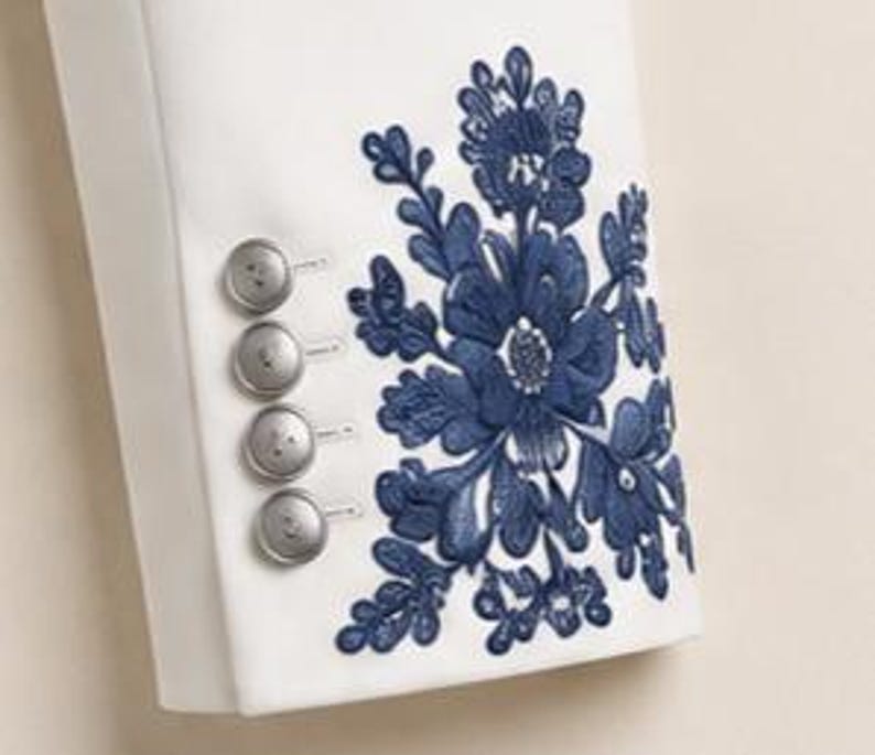 May include: A white sleeve featuring a vertical row of four round silver buttons and a large, intricate dark blue floral embroidery pattern. The embroidery consists of detailed petals and leaves, positioned next to the buttons on the fabric.