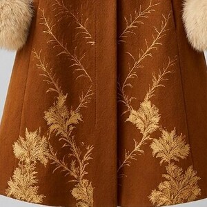 May include: A brown coat featuring a gold-colored botanical pattern of fern-like leaves extending upward from the hem. The garment includes long sleeves finished with thick, fluffy beige faux fur cuffs at the ends of the sleeves.