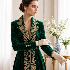 May include: A long-sleeved, floor-length emerald green velvet coat featuring intricate gold floral embroidery along the lapels and front closure. The coat includes a row of gold buttons down the center and on the cuffs, paired with cream-colored gloves.
