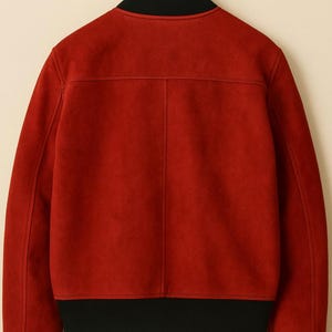 May include: A red bomber jacket with a black collar, cuffs, and hem. The jacket is hanging on a wooden hanger. The back of the jacket is visible, showing the seams and texture of the fabric.