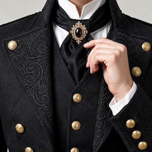 May include: A black brocade jacket featuring gold-toned embossed buttons, a white collared shirt, and a black satin cravat secured with an oval-shaped gold brooch containing a central black stone. The jacket has intricate floral patterns.