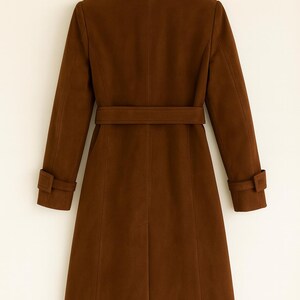 May include: A brown coat with a belt and long sleeves. The coat is hanging on a wooden hanger. The coat has a classic design with a collar and buttoned cuffs.
