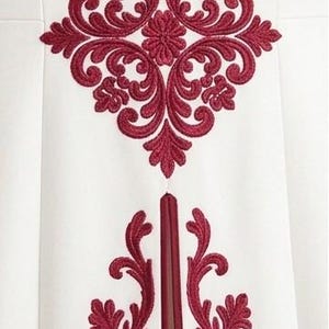 May include: A white garment featuring a prominent, symmetrical dark red embroidered damask pattern centered at the top, with two matching vertical red embroidered floral motifs extending downwards on either side of a central vertical seam.