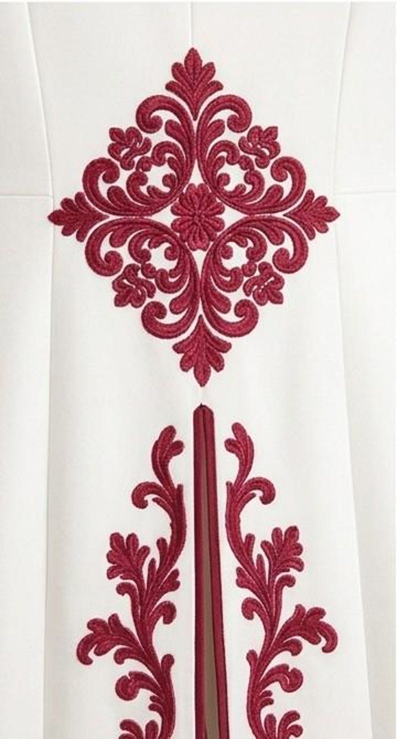 May include: A white garment featuring a prominent, symmetrical dark red embroidered damask pattern centered at the top, with two matching vertical red embroidered floral motifs extending downwards on either side of a central vertical seam.