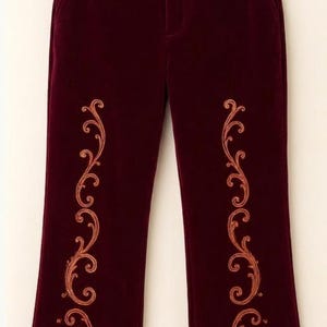 Pu&ograve; includere: Pantaloni in velluto bordeaux scuro con un motivo a volute ricamato color rame su ogni gamba e un bordo spesso in pelliccia sintetica color marrone chiaro sull'orlo inferiore di ogni gamba, appesi a una gruccia di legno.