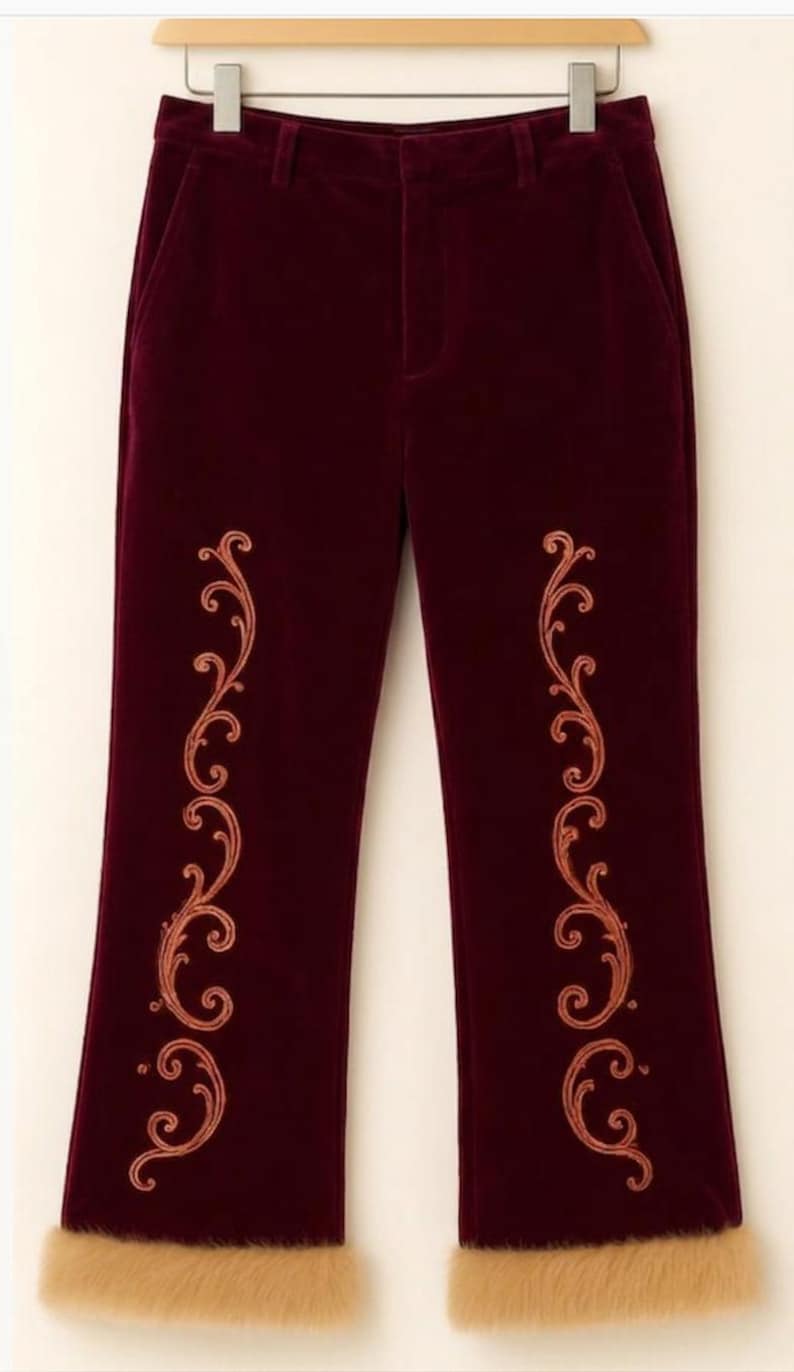 Pu&ograve; includere: Pantaloni in velluto bordeaux scuro con un motivo a volute ricamato color rame su ogni gamba e un bordo spesso in pelliccia sintetica color marrone chiaro sull'orlo inferiore di ogni gamba, appesi a una gruccia di legno.