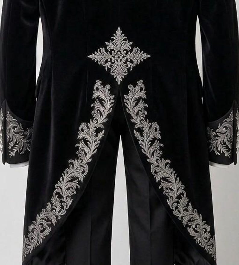 May include: A black velvet tailcoat featuring intricate silver embroidery along the cuffs and the back panels. The design includes a prominent floral-inspired motif centered at the waist and scrolling leaf patterns lining the coat tails.