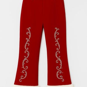 Pu&ograve; includere: Pantaloni rosso scuro con gamba a zampa e bordo in pelliccia sintetica bianca sugli orli. La parte anteriore di ogni gamba &egrave; decorata con un intricato motivo ricamato a filigrana color chiaro che si estende verticalmente.