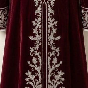 Pu&ograve; includere: Un cappotto in velluto bordeaux intenso caratterizzato da un intricato ricamo argentato che corre verticalmente lungo il centro e l'orlo. Le maniche lunghe sono rifinite con polsini ricamati in argento abbinati, creando un design dettagliato.