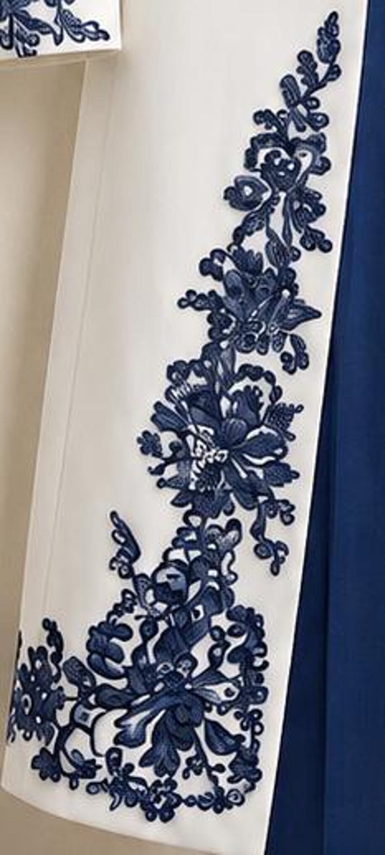 May include: A white garment panel featuring an intricate dark blue floral embroidery pattern running vertically along the edge. The embroidery consists of detailed leaves and blossoms, contrasting sharply against the smooth white fabric base.