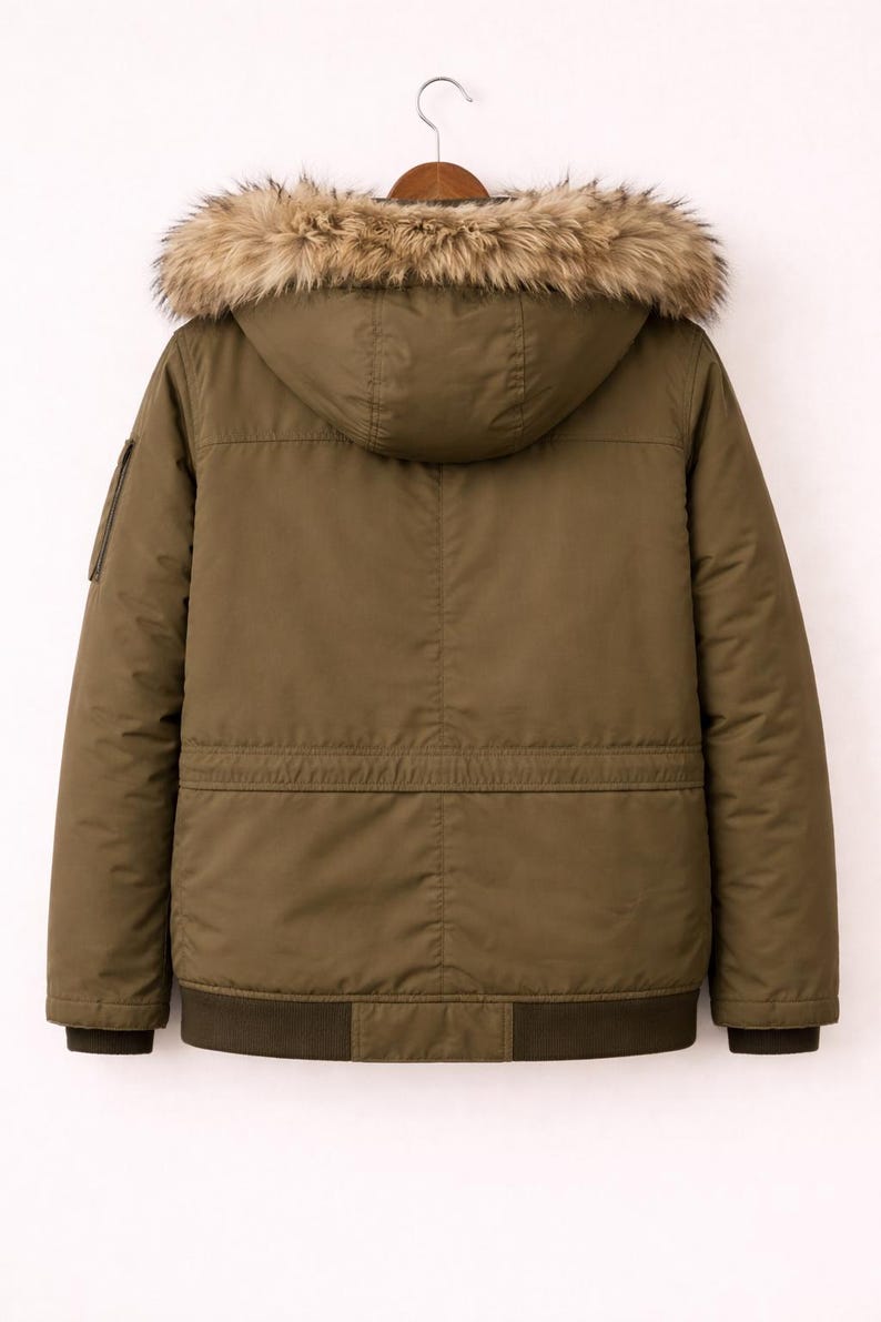 May include: Olive green winter jacket with a hood and faux fur trim. The jacket has a bomber-style bottom and a small pocket on the left sleeve. The jacket is hanging on a wooden hanger.