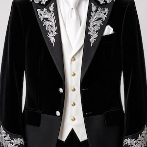 May include: A black velvet tailcoat featuring silver floral embroidery on the lapels and cuffs, paired with a white waistcoat with gold buttons, a white dress shirt, and a white necktie. The jacket has a high collar and a pocket square.