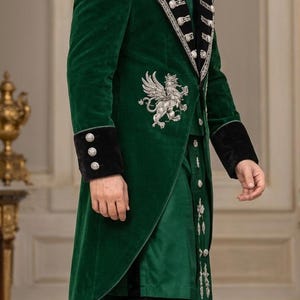 May include: A deep green velvet long coat featuring black velvet lapels and cuffs, adorned with silver metallic embroidery of a griffin on the chest and silver buttons. The coat is worn over a matching green tunic and solid black trousers.