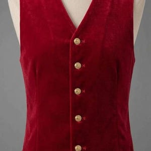 May include: A sleeveless vest made of deep red velvet fabric. The garment features a V-neckline and a front button closure with five round, metallic gold-toned buttons. The hem of the vest is pointed at the center front.