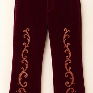 May include: Deep burgundy velvet trousers featuring a vertical copper-colored embroidered scrollwork pattern on each leg and a thick, light tan faux fur trim along the bottom hem of each pant leg, suspended from a wooden clothing hanger.