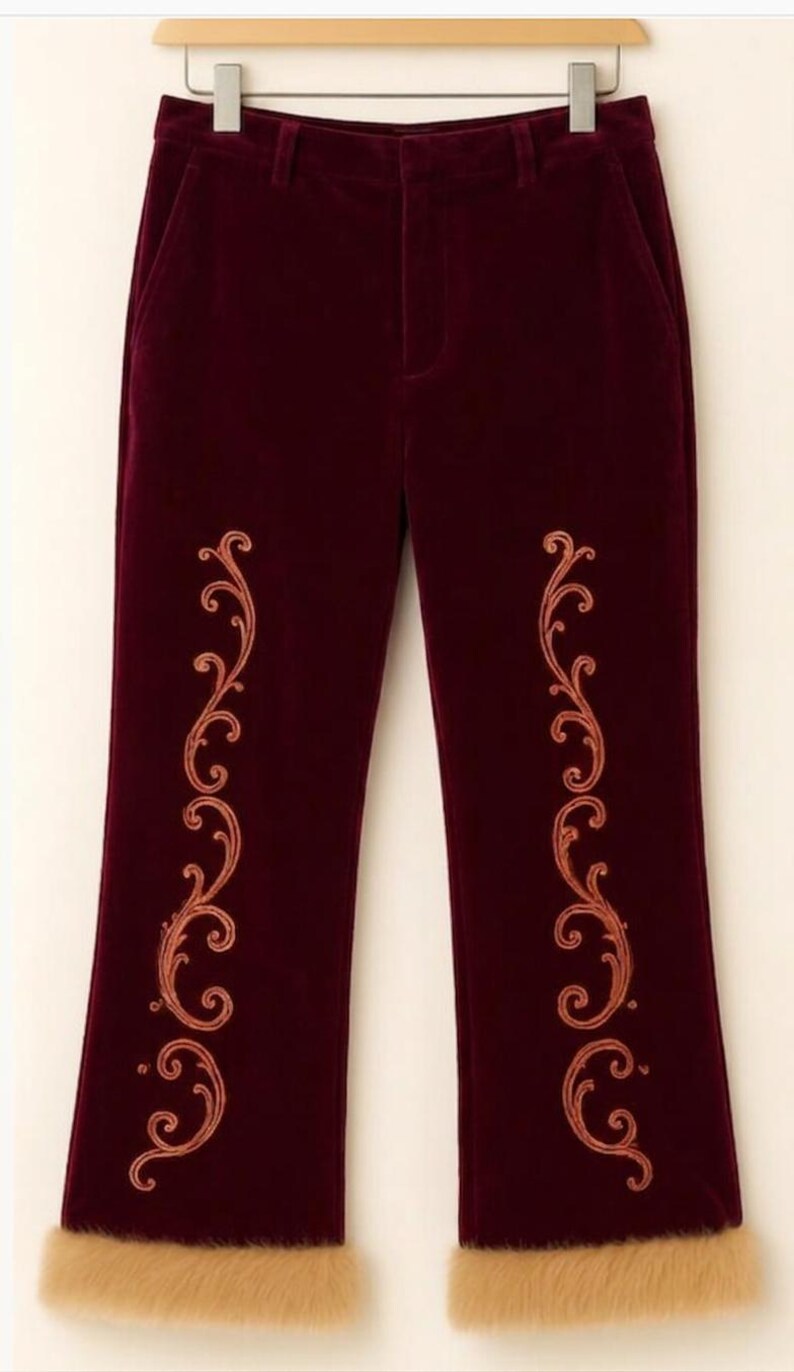May include: Deep burgundy velvet trousers featuring a vertical copper-colored embroidered scrollwork pattern on each leg and a thick, light tan faux fur trim along the bottom hem of each pant leg, suspended from a wooden clothing hanger.