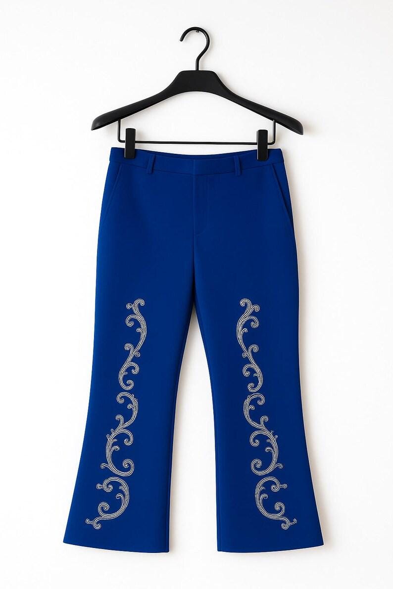May include: Royal blue trousers featuring a flared leg silhouette and a waistband with belt loops. The front of each pant leg is decorated with a vertical, symmetrical silver-toned filigree scroll pattern embroidered along the center.