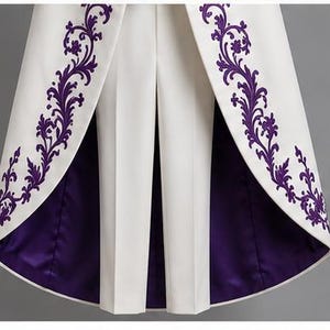 May include: A white coat with a high-low hemline featuring deep purple floral embroidery along the front edges. The interior lining of the coat is a solid deep purple satin, and it is worn over a pair of matching white tailored trousers.
