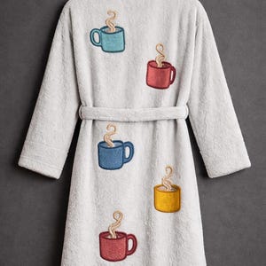 Puede incluir: Una bata de ba&ntilde;o gris claro y afelpada con cintur&oacute;n a juego. La bata est&aacute; decorada con tazas de caf&eacute; bordadas en azul, rojo, amarillo y verde azulado, cada una con vapor saliendo. La bata est&aacute; colgada de un gancho negro.