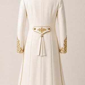 May include: A long, off-white coat featuring a high collar, long sleeves, and a structured back panel. The garment is adorned with intricate gold embroidery on the collar, cuffs, and a horizontal waist tab, complemented by gold piping along the edges.