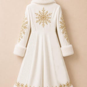 May include: A long white coat featuring a plush white faux fur collar and sleeve cuffs. The back of the coat is decorated with a large gold embroidered snowflake design, with additional gold snowflake patterns on the sleeves and hemline.