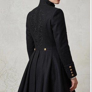 May include: A black coat featuring a high collar, long sleeves with four gold buttons on each cuff, and a pleated skirt. The back of the coat is decorated with intricate black embroidery and two gold buttons at the waistline.
